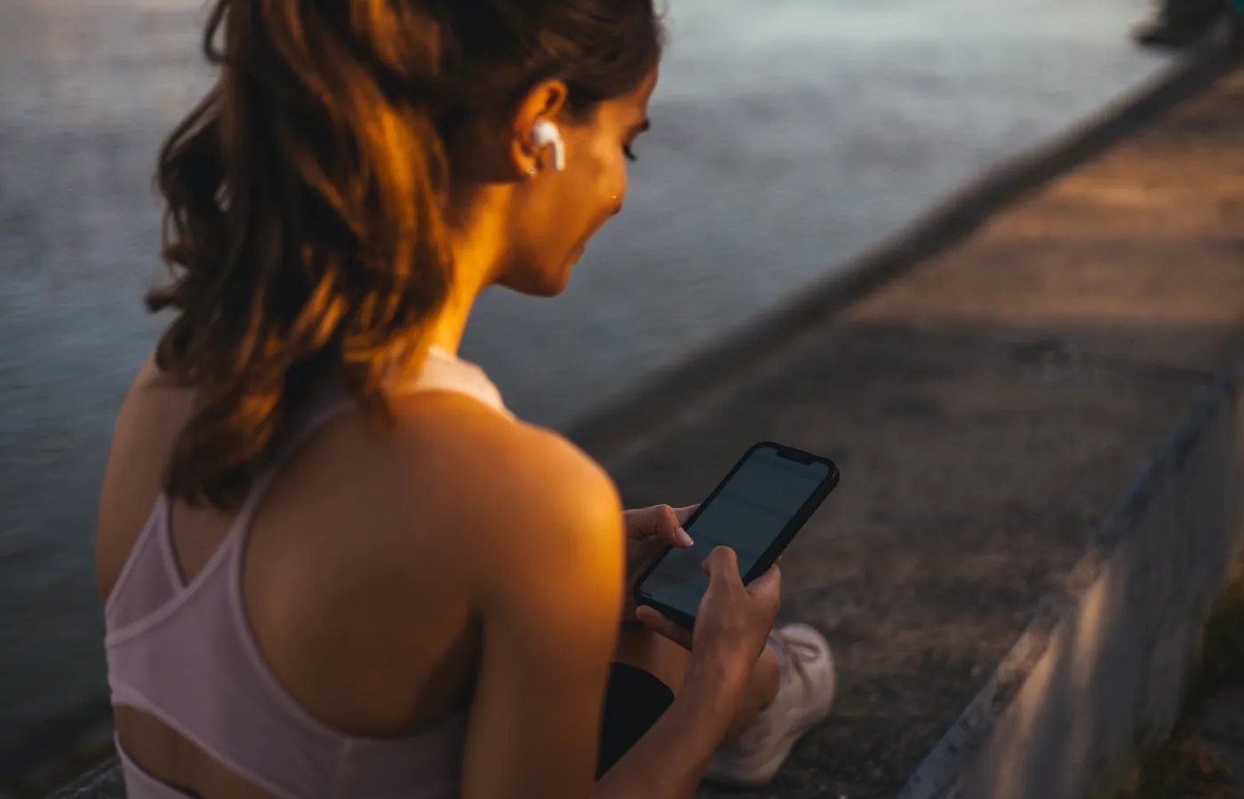 A woman sitting on a sea wall at sunset, looking at her phone after a run.